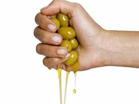 A hand squeezing a cluster of green olives, with olive oil dripping from them. - Olive Oil Times