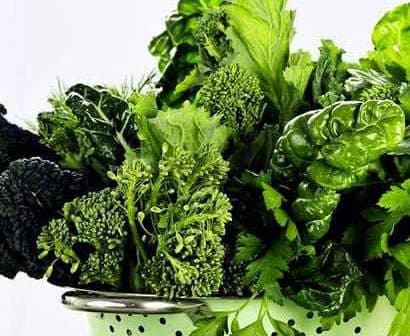 A colander filled with various green leafy vegetables including kale, spinach, and herbs. - Olive Oil Times
