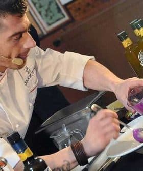 Chef in a white coat preparing a dish with olive oil and colorful ingredients on a countertop. - Olive Oil Times