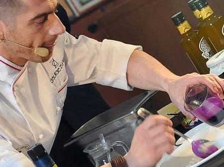 Chef in a white coat preparing a dish with olive oil and colorful ingredients on a countertop. - Olive Oil Times