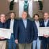 Five individuals posing together at an event, holding certificates in front of a sculpture backdrop. - Olive Oil Times