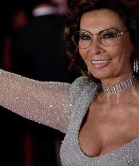 Woman wearing a sparkling silver dress and glasses, smiling and waving at an event. - Olive Oil Times