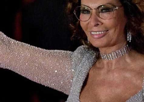 Woman wearing a sparkling silver dress and glasses, smiling and waving at an event. - Olive Oil Times
