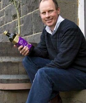 Man sitting on a step holding a bottle of olive oil with a tree in the background. - Olive Oil Times