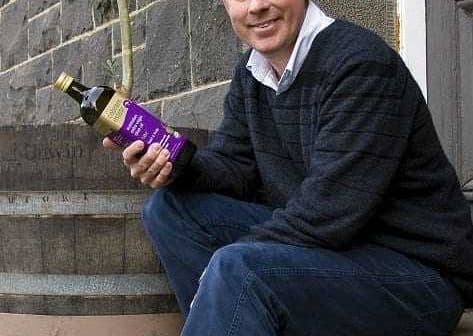 Man sitting on a step holding a bottle of olive oil with a tree in the background. - Olive Oil Times