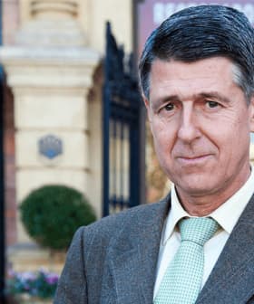 Man wearing a brown suit and a green tie, standing outdoors near a building entrance. - Olive Oil Times