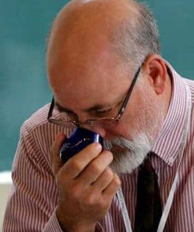 A man with glasses and a beard smelling a blue container in a classroom setting. - Olive Oil Times