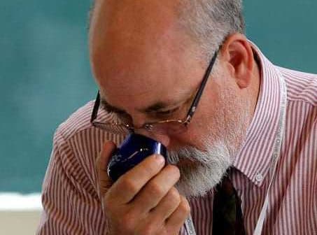 A man with glasses and a beard smelling a blue container in a classroom setting. - Olive Oil Times