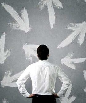 Individual in a white shirt standing with hands on hips, facing a wall covered in white arrow patterns. - Olive Oil Times