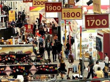 View of aisles at a food trade show with signage indicating aisle numbers and various booths. - Olive Oil Times
