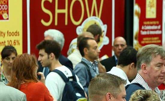 Crowd of attendees at a food show event with a large red banner in the background. - Olive Oil Times