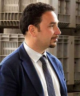 Profile view of a man wearing a suit and tie, standing in front of storage containers. - Olive Oil Times