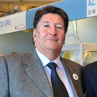 A man wearing a grey suit and a blue tie, standing in an indoor setting with a neutral expression. - Olive Oil Times