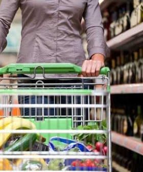 Person pushing a shopping cart filled with groceries in a supermarket aisle with shelves of bottles. - Olive Oil Times