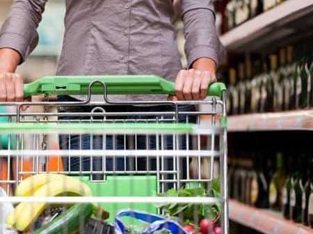 Person pushing a shopping cart filled with groceries in a supermarket aisle with shelves of bottles. - Olive Oil Times