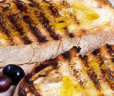Two slices of grilled bread topped with olive oil and grill marks on a wooden board. - Olive Oil Times