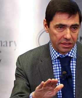 Man in a gray suit with a blue checkered shirt speaking during a presentation. - Olive Oil Times