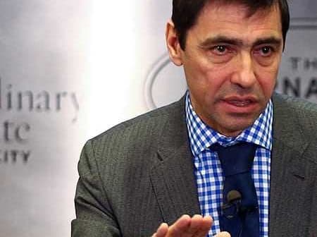 Man in a gray suit with a blue checkered shirt speaking during a presentation. - Olive Oil Times