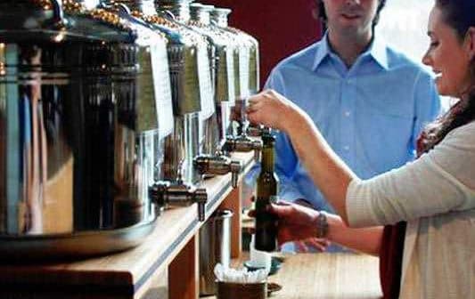 A woman dispensing oil from a large metal container at a service station while a man observes. - Olive Oil Times