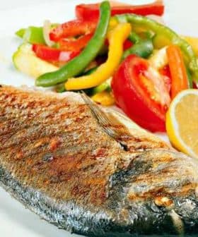 Grilled fish served with a side of colorful sautéed vegetables and lemon slices on a white plate. - Olive Oil Times