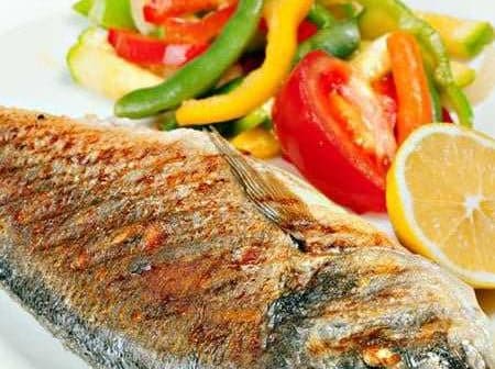 Grilled fish served with a side of colorful sautéed vegetables and lemon slices on a white plate. - Olive Oil Times