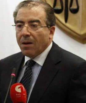 A man in a suit speaking at a podium with microphones and a backdrop featuring scales of justice. - Olive Oil Times