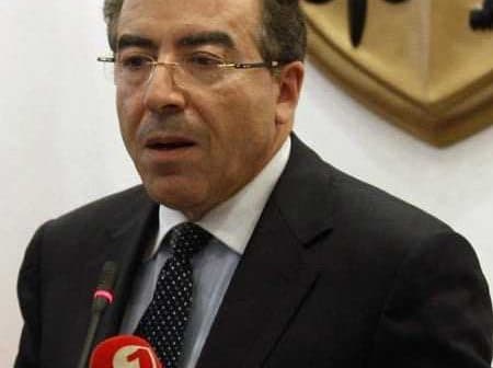 A man in a suit speaking at a podium with microphones and a backdrop featuring scales of justice. - Olive Oil Times