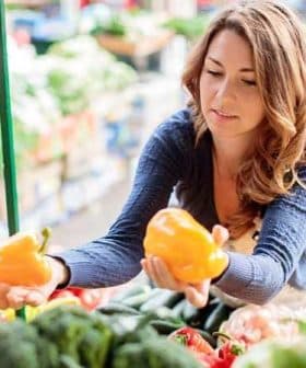 Woman examining yellow bell peppers while shopping at a market stall filled with vegetables. - Olive Oil Times