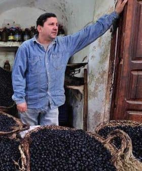 A man in a blue shirt standing next to large baskets filled with black olives. - Olive Oil Times