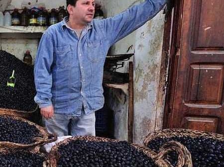A man in a blue shirt standing next to large baskets filled with black olives. - Olive Oil Times