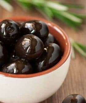 A small bowl filled with black olives placed on a wooden surface. - Olive Oil Times