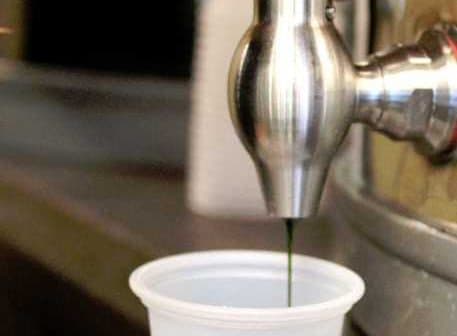 A hand operating a metal oil dispenser to fill a clear plastic cup with dark liquid. - Olive Oil Times
