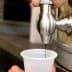 A hand operating a metal oil dispenser to fill a clear plastic cup with dark liquid. - Olive Oil Times