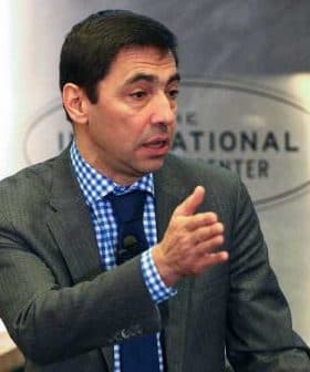 Man in a gray suit and blue checkered shirt gesturing while speaking at a conference. - Olive Oil Times