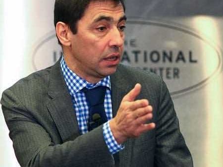 Man in a gray suit and blue checkered shirt gesturing while speaking at a conference. - Olive Oil Times