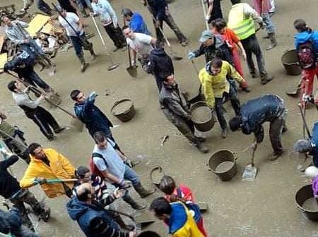 A large group of individuals using shovels and buckets in a flooded area, wearing various clothing and engaged in cleanup efforts. - Olive Oil Times