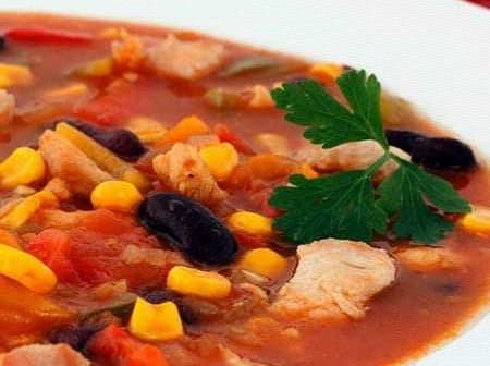 Bowl of chicken and vegetable soup with corn, beans, and herbs on a white plate. - Olive Oil Times