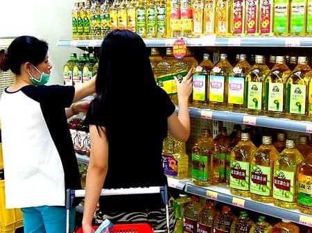 Two individuals shopping for cooking oils on a supermarket shelf filled with various oil bottles. - Olive Oil Times