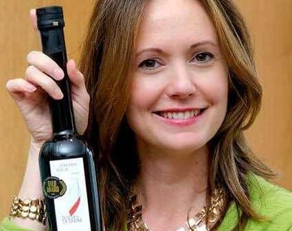 Woman smiling while holding a bottle of olive oil in her right hand. - Olive Oil Times