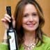 Woman smiling while holding a bottle of olive oil in her right hand. - Olive Oil Times