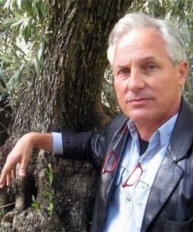 A man in a leather jacket leaning against the trunk of an olive tree with glasses in hand. - Olive Oil Times