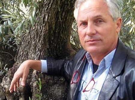 A man in a leather jacket leaning against the trunk of an olive tree with glasses in hand. - Olive Oil Times