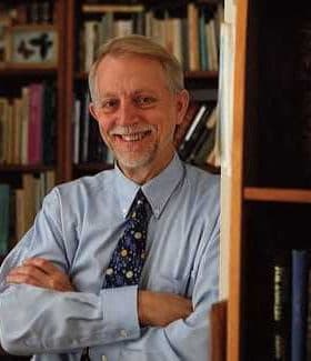A man with gray hair and glasses smiling while standing in front of bookshelves filled with books. - Olive Oil Times