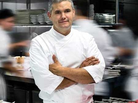 A chef standing with arms crossed in a busy professional kitchen environment. - Olive Oil Times