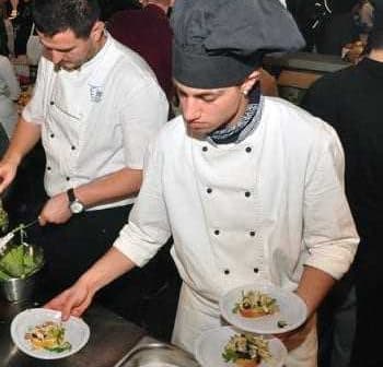 Two chefs in white uniforms plating food in a busy kitchen environment. - Olive Oil Times