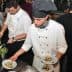Two chefs in white uniforms plating food in a busy kitchen environment. - Olive Oil Times