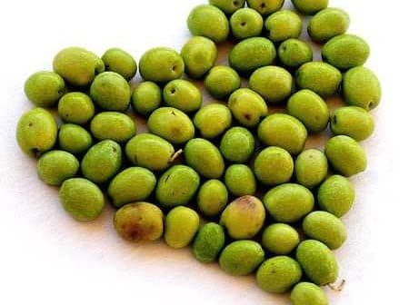 A collection of green olives arranged in the shape of a heart on a white background. - Olive Oil Times