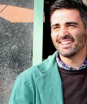 Man wearing a green jacket and a dark sweater, smiling while standing outdoors near a window. - Olive Oil Times