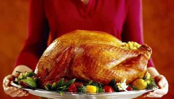 A person holding a large roasted turkey on a silver serving platter with garnishes. - Olive Oil Times