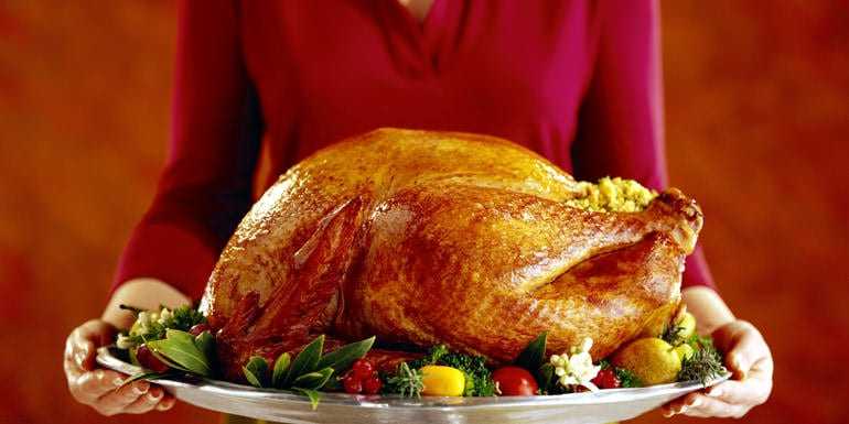 A person holding a large roasted turkey on a silver serving platter with garnishes. - Olive Oil Times